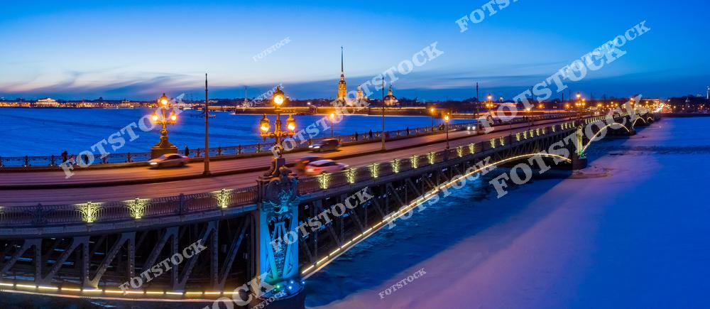 На фотографии изображён Троицкий мост, освещённый вечерними огнями. Под мостом видна замёрзшая поверхность реки Невы, а на заднем плане выделяется Петропавловская крепость. Освещение и синие оттенки зимнего заката создают атмосферу спокойствия и величия. Это одно из самых узнаваемых мест Санкт-Петербурга.